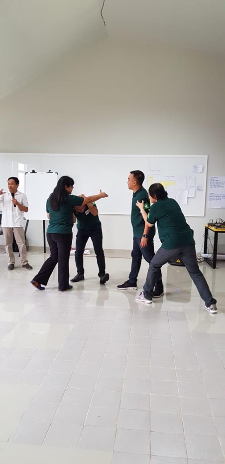 A stack of books and a green recycling bin sit on a tiled floor, with a tan couch and bookshelf in the background.